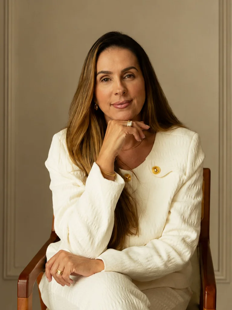 Dra. Adriana Mestre sentada, de blazer claro, em ambiente com painéis em tom neutro.